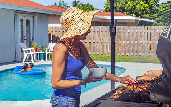 Woman cooking on a propane grill in a backyard with a swimming pool in the background